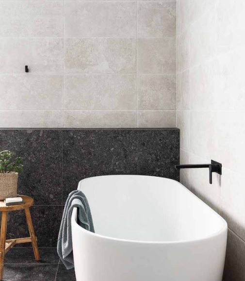 Bathroom with a white freestanding bathtub, gray wall tiles, and a dark gray stone backsplash. A wooden stool and a black faucet are also visible.