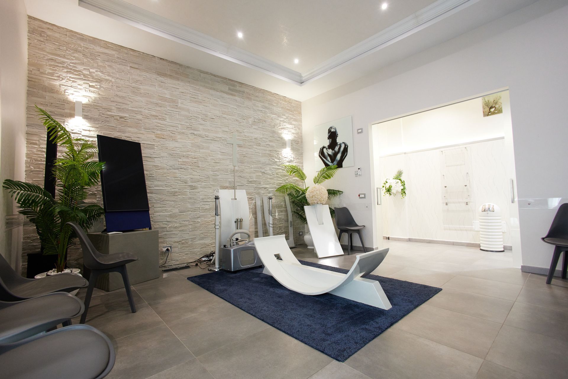 Moderna sala d'attesa con decorazioni bianche e grigie; una sedia bianca curva, piante e un tappeto blu scuro.