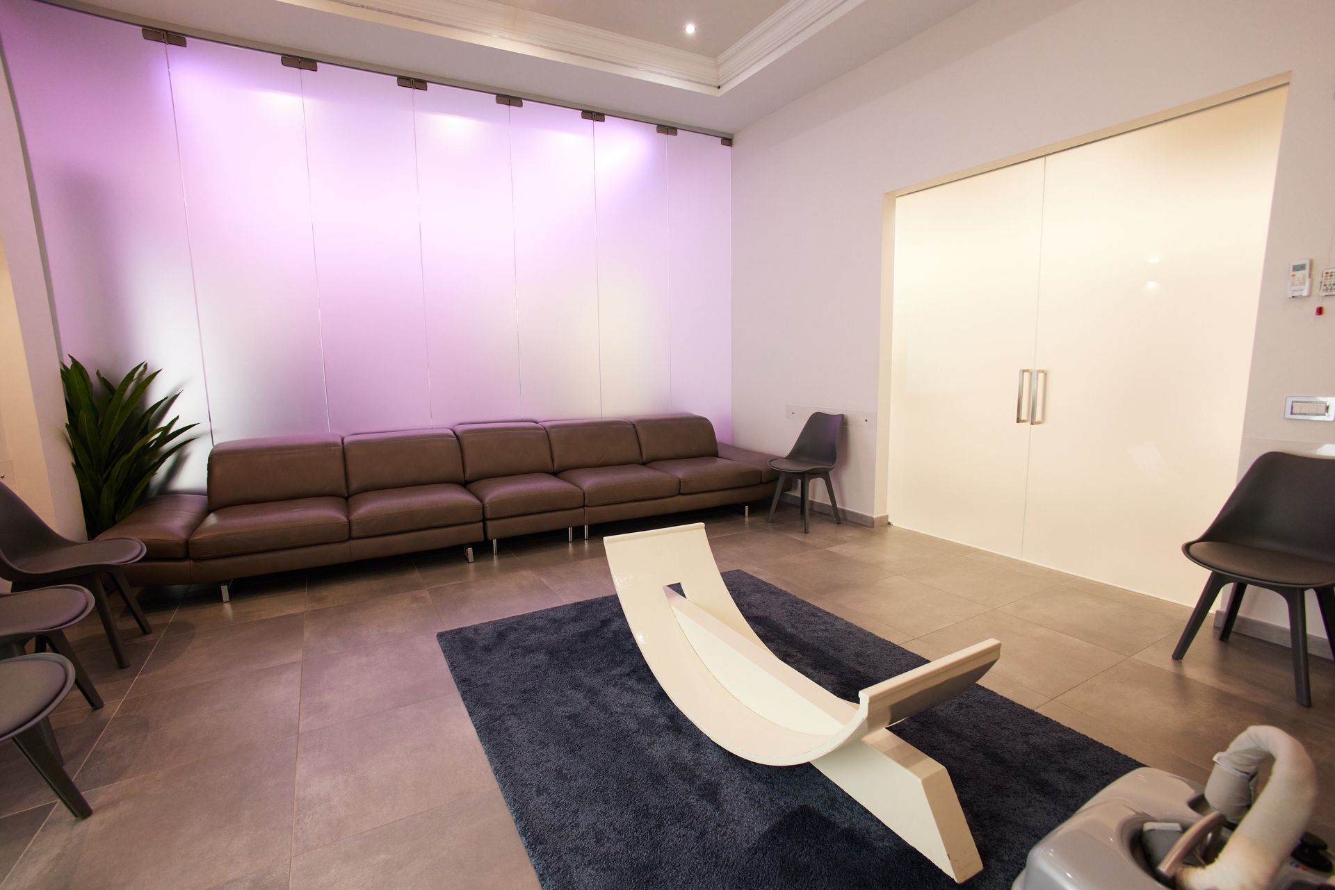 Sala d'attesa con divano marrone, sedie e una balance board in legno su un tappeto blu.