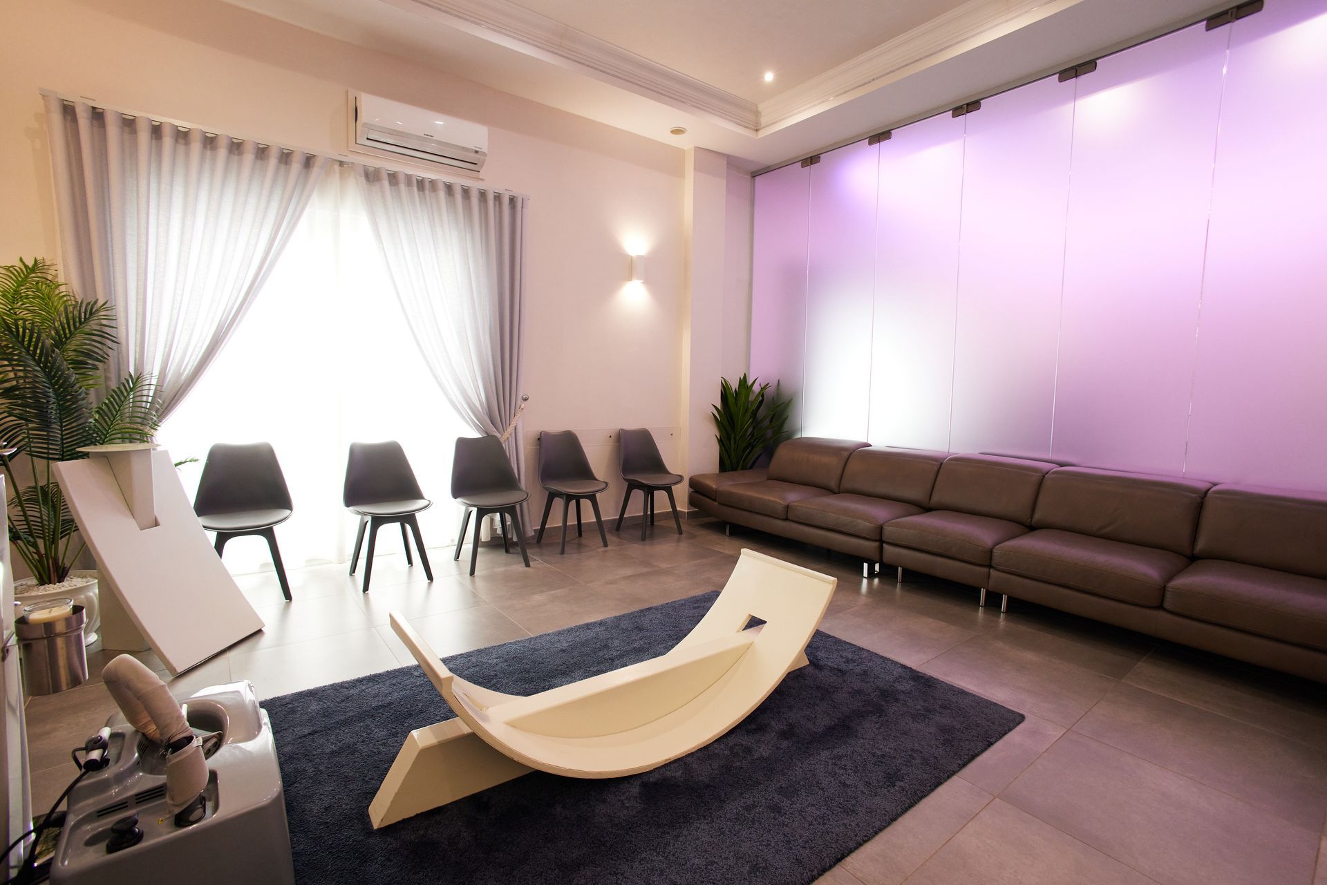 Sala d'attesa con sedie, un lungo divano marrone, una finestra, piante e una panca di legno su un tappeto.