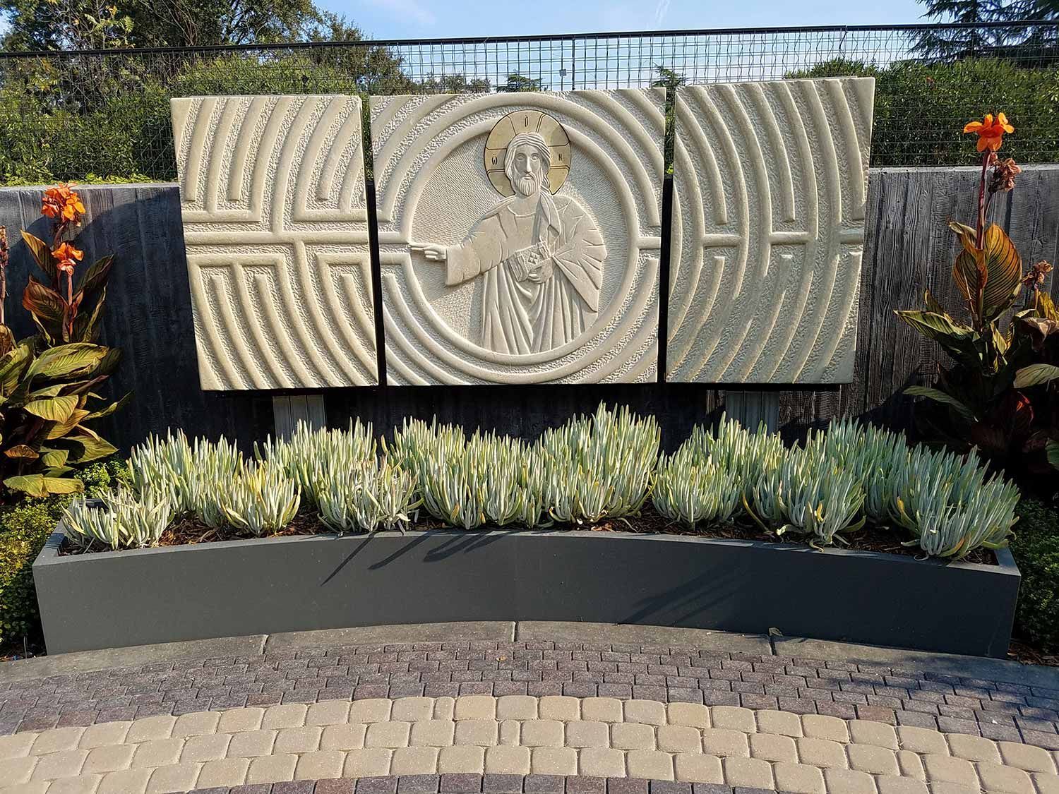 Concrete walkway bordered by brown mulch and green and yellow bushes, leading toward buildings.