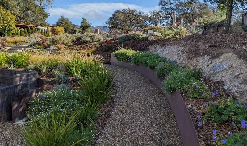 Gravel pathway winds through a garden with greenery and planted beds. Sunny day.