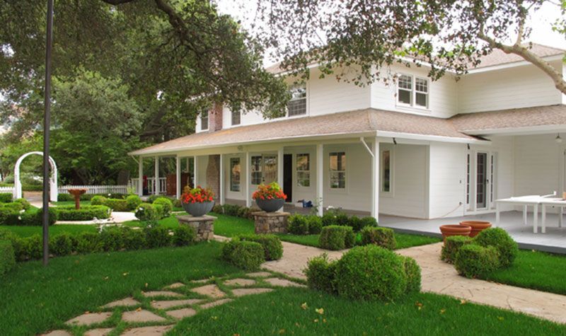 White house with a wrap-around porch and manicured lawn with hedges and stone pathway.