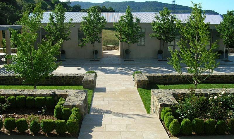 Stone path leads to a building with trees, flanked by garden beds, in a rural setting.
