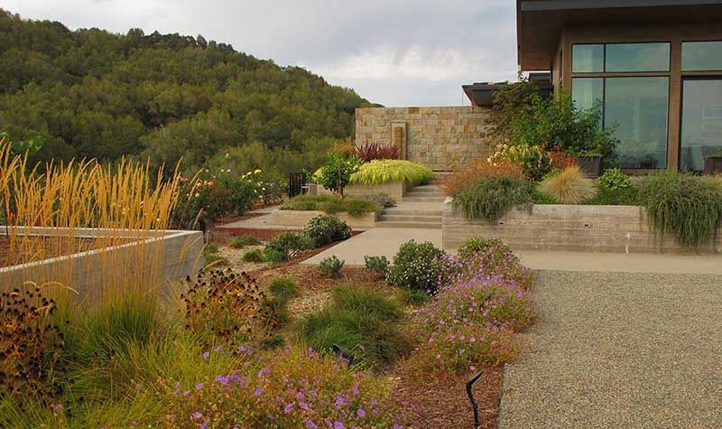 Landscaped garden with gravel path, stone walls, and tiered planting beds with colorful flowers against a hillside.
