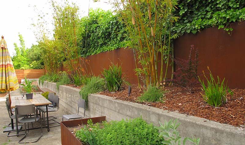A backyard patio with a raised garden bed and a brown metal fence. Bamboo and other plants grow along the fence.