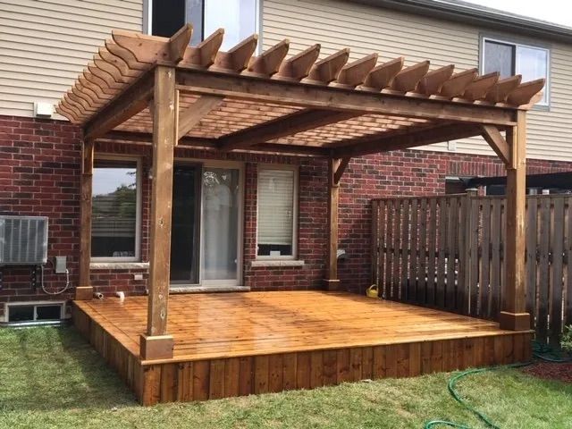 A wooden pergola is sitting on top of a wooden deck in front of a brick house.