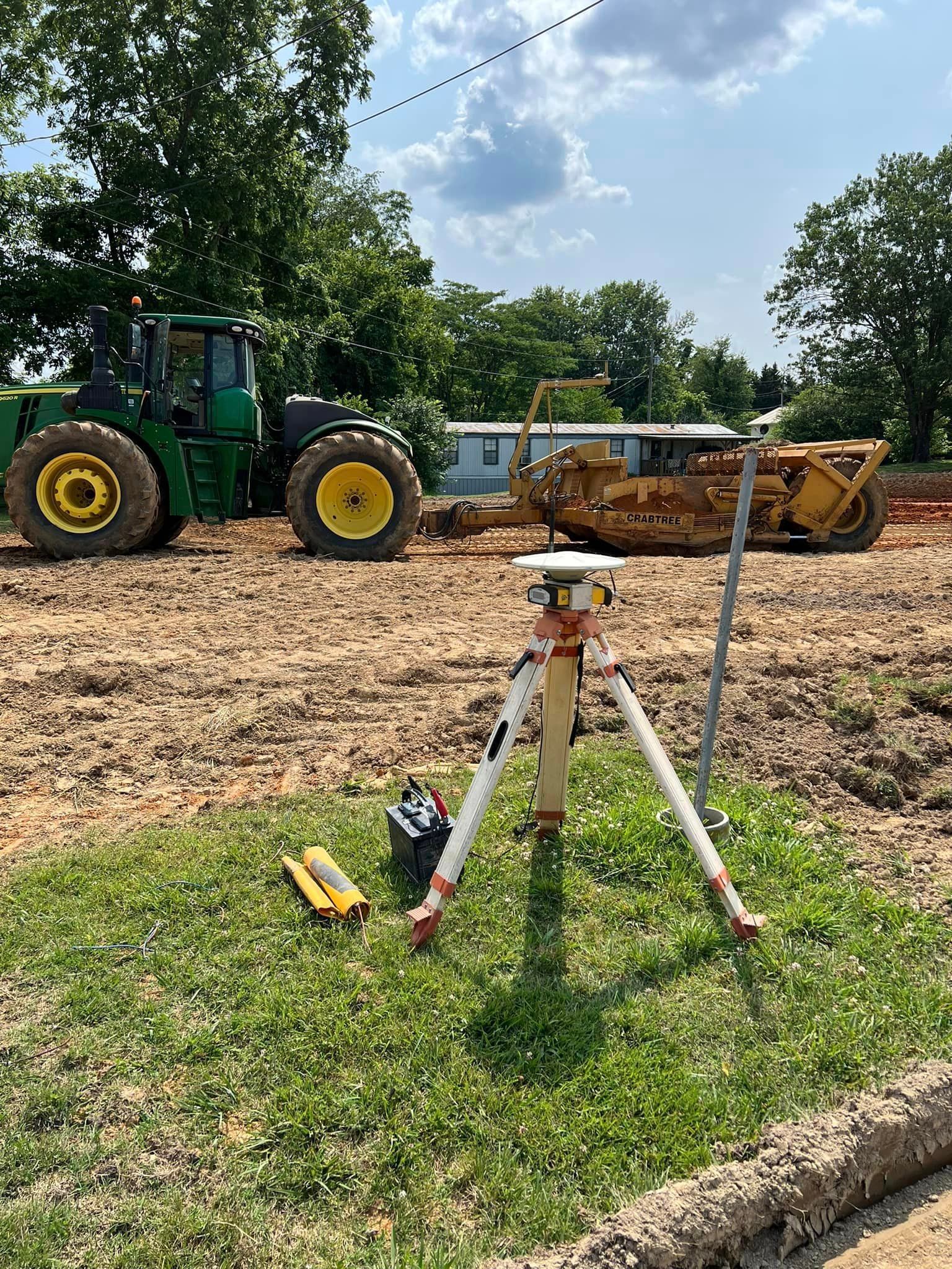 A tripod is sitting in the grass next to a tractor on a construction site.