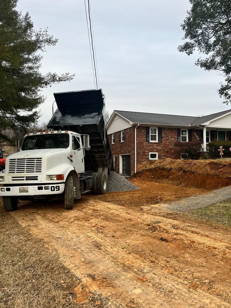 A dump truck is driving down a dirt road in front of a house.
