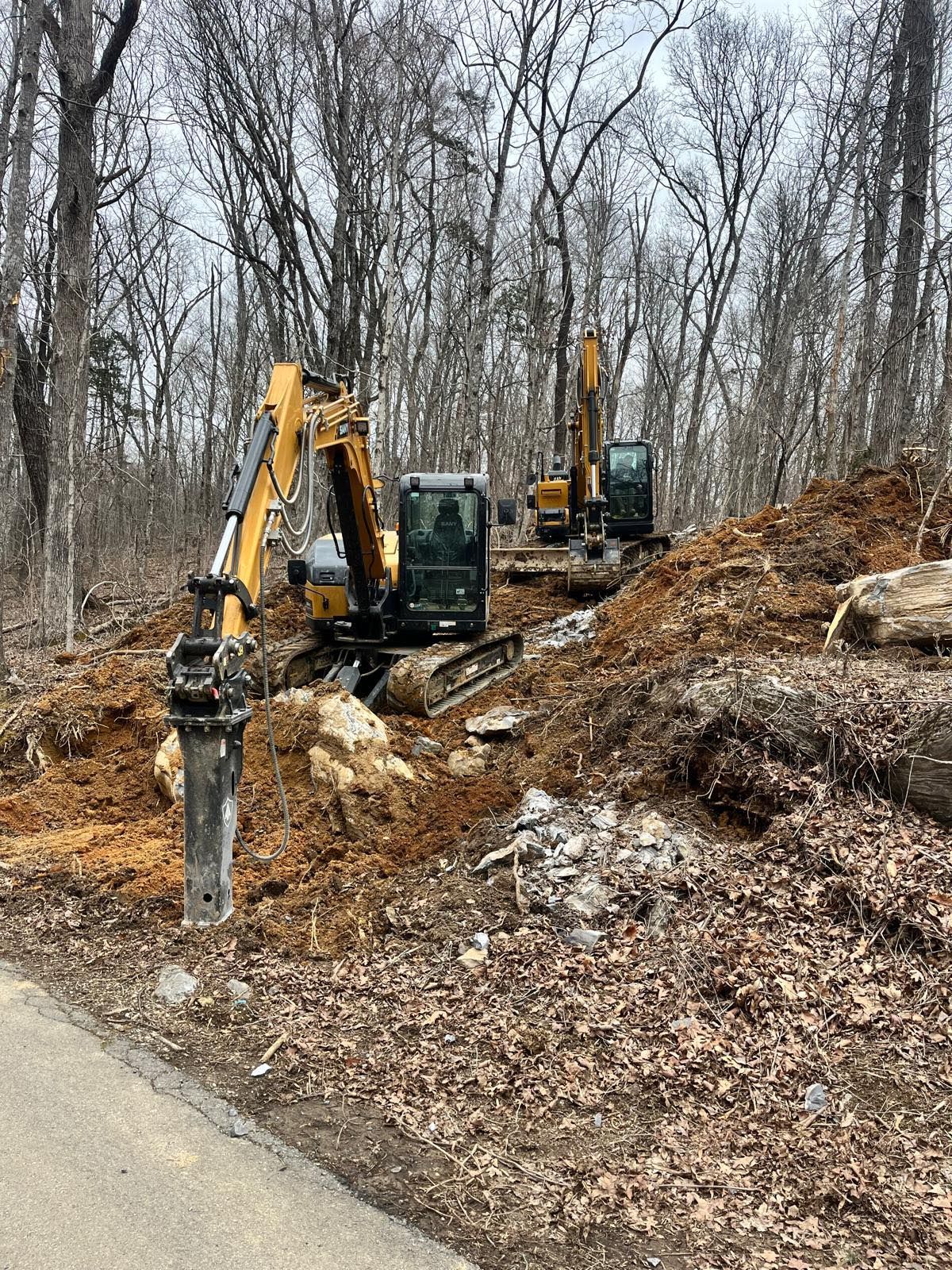Two excavators are working on a construction site in the woods.