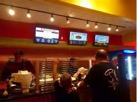 Restaurant counter with screens, staff, and customers; red wall, track lighting, and food displays visible.