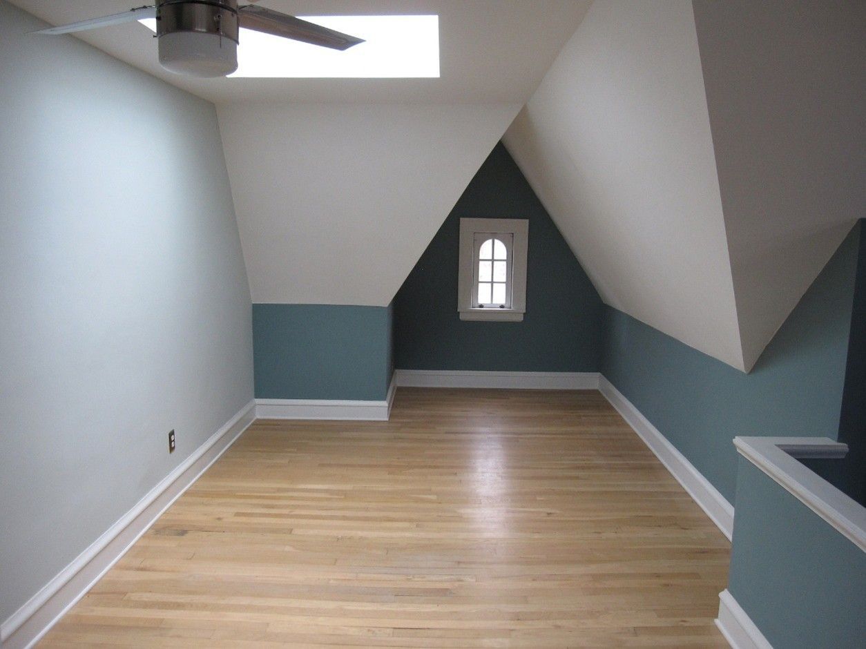 Empty attic room with wood floor, blue and white walls, small window, and ceiling fan.