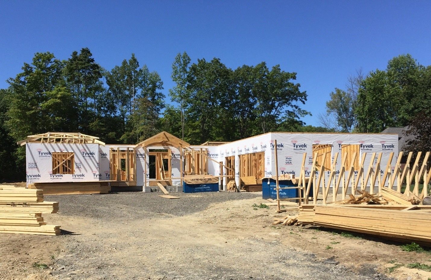 A house under construction. Wood framing and sheathing visible. Clear blue sky and trees in the background.