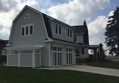 A light grey two-story house with a Dutch gambrel roof and two garage doors. A white pergola and driveway are present.