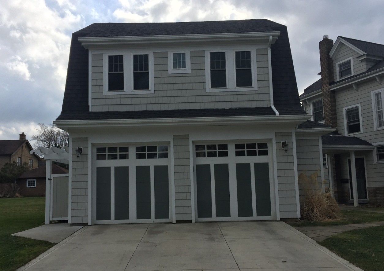 Two-car garage with a second-story living space; light gray siding, dark roof, two garage doors, and several windows.