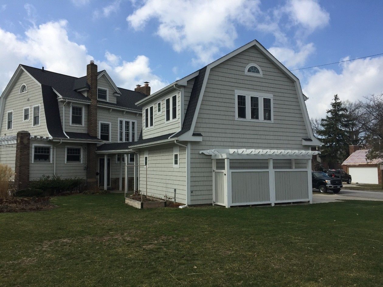 Two-story house with light gray siding, dark roof, and a detached garage on a grassy lawn under a partly cloudy sky.