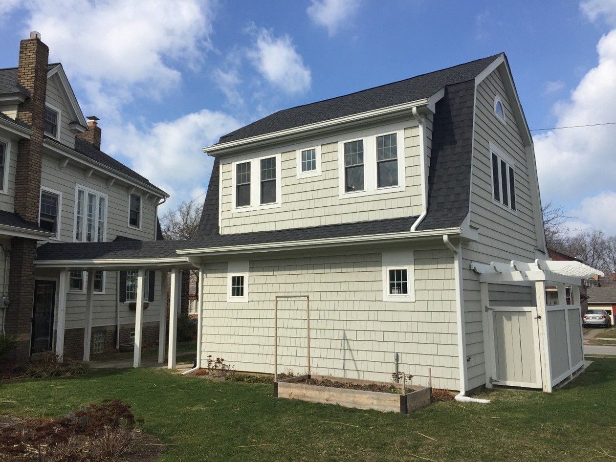 Two-story gray building with a unique roof design, attached to a light-colored house. Green grass and blue sky.