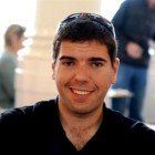 Smiling man with dark hair wearing sunglasses on his head, in a bright room. Two blurred figures are in the background.