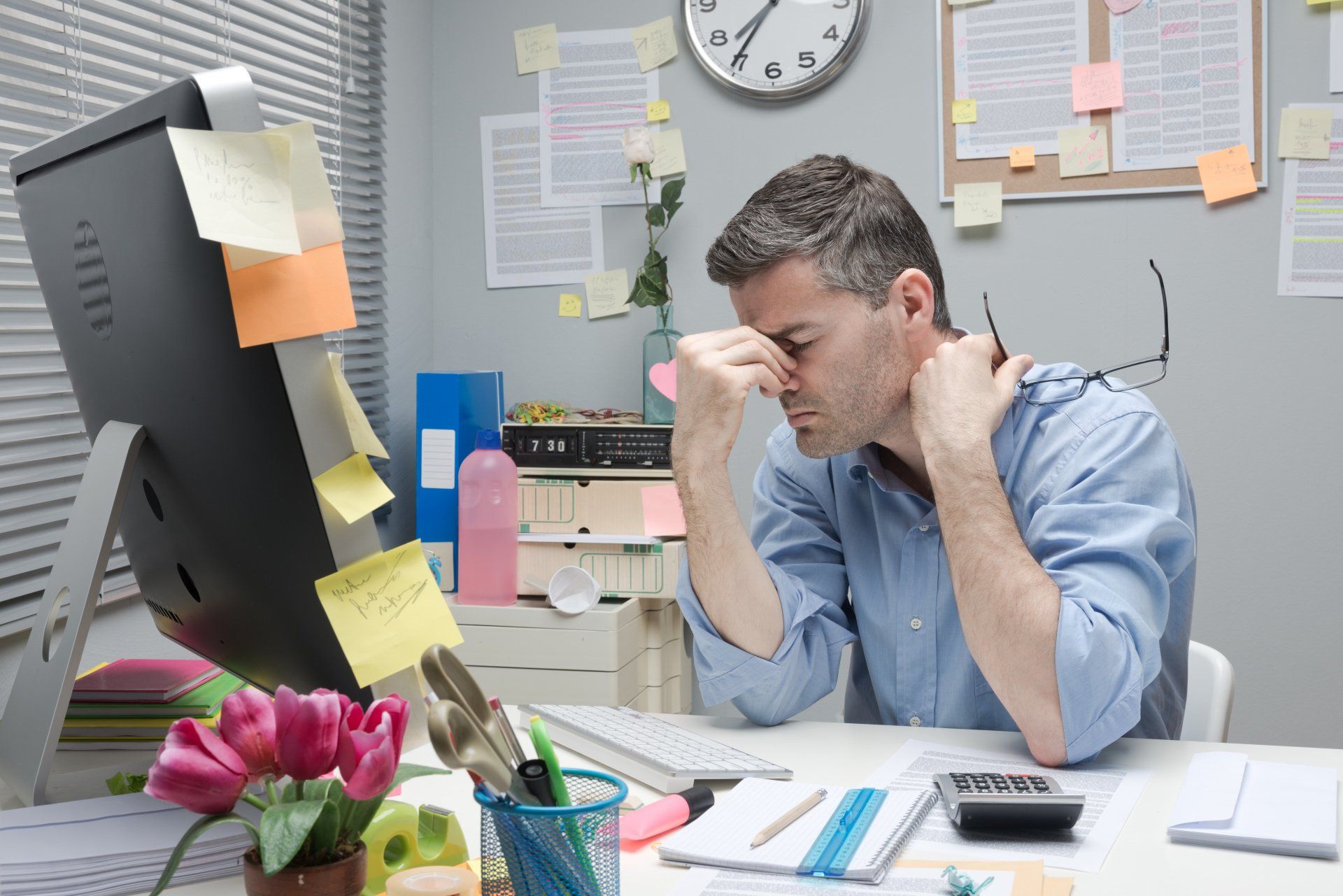 Man at a cluttered desk, stressed, holding his face, surrounded by sticky notes and a computer.