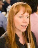 Woman with red hair and bangs, in a black top, talking.  She is in a room with others.