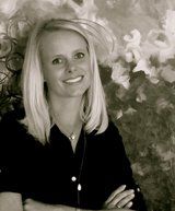 Blonde woman with arms crossed smiling, wearing a black shirt, in front of an abstract floral painting.