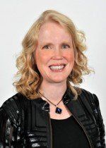 Blonde woman with curly hair smiles, wearing a black jacket and a necklace with a blue pendant.