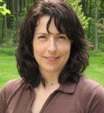 Woman with brown hair, smiling, wearing a brown shirt, outdoors with green grass and trees in the background.