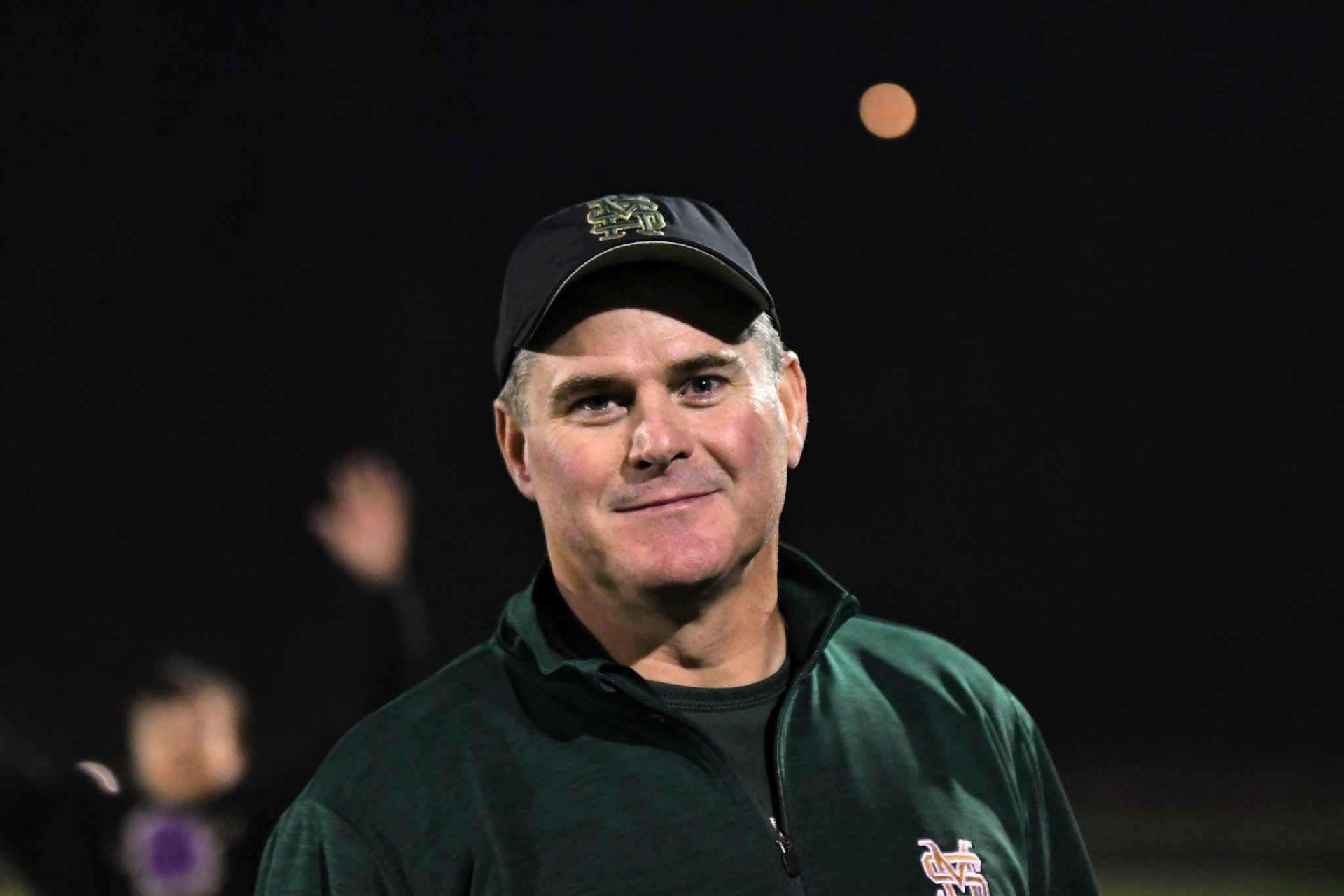A man in a green jacket and baseball cap smiles outdoors at night, a ball floating above him.