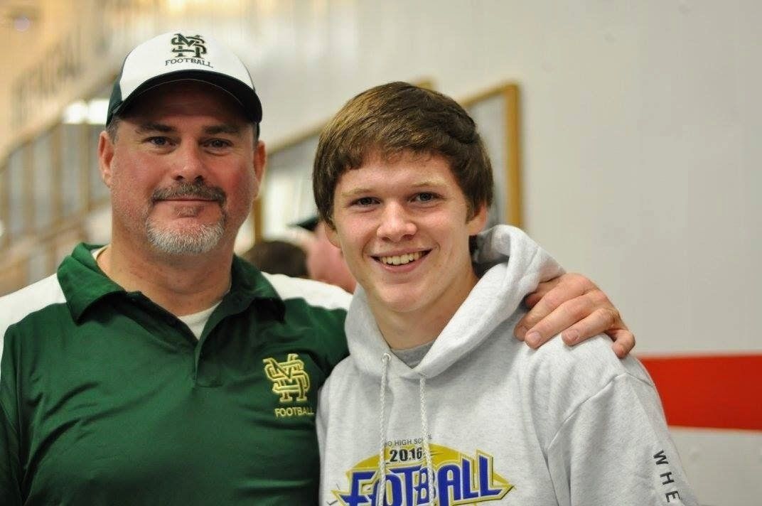 Man with a beard and a baseball cap puts arm around a smiling teenage boy wearing a hoodie. They stand indoors.