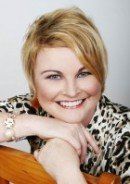 Woman with short, blonde hair smiles, resting arms on a wooden chair, wearing a leopard print top.