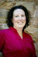 Woman with curly dark hair smiles at the camera, wearing a magenta top against a stone wall backdrop.
