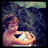 Woman with short red hair, wearing sunglasses, in a white dress, sitting by dark rocky cliffs.