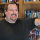 Man with a beard smiling, leaning against a bookshelf filled with books and DVDs.