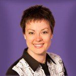 Woman with short brown hair, smiling, wearing a black and white collared shirt, in front of a purple background.