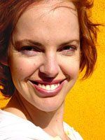 Woman with red hair smiles broadly against a yellow background. She wears a white top and has freckles.
