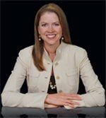 Woman in a light jacket smiles, resting arms on a dark surface against a black background.
