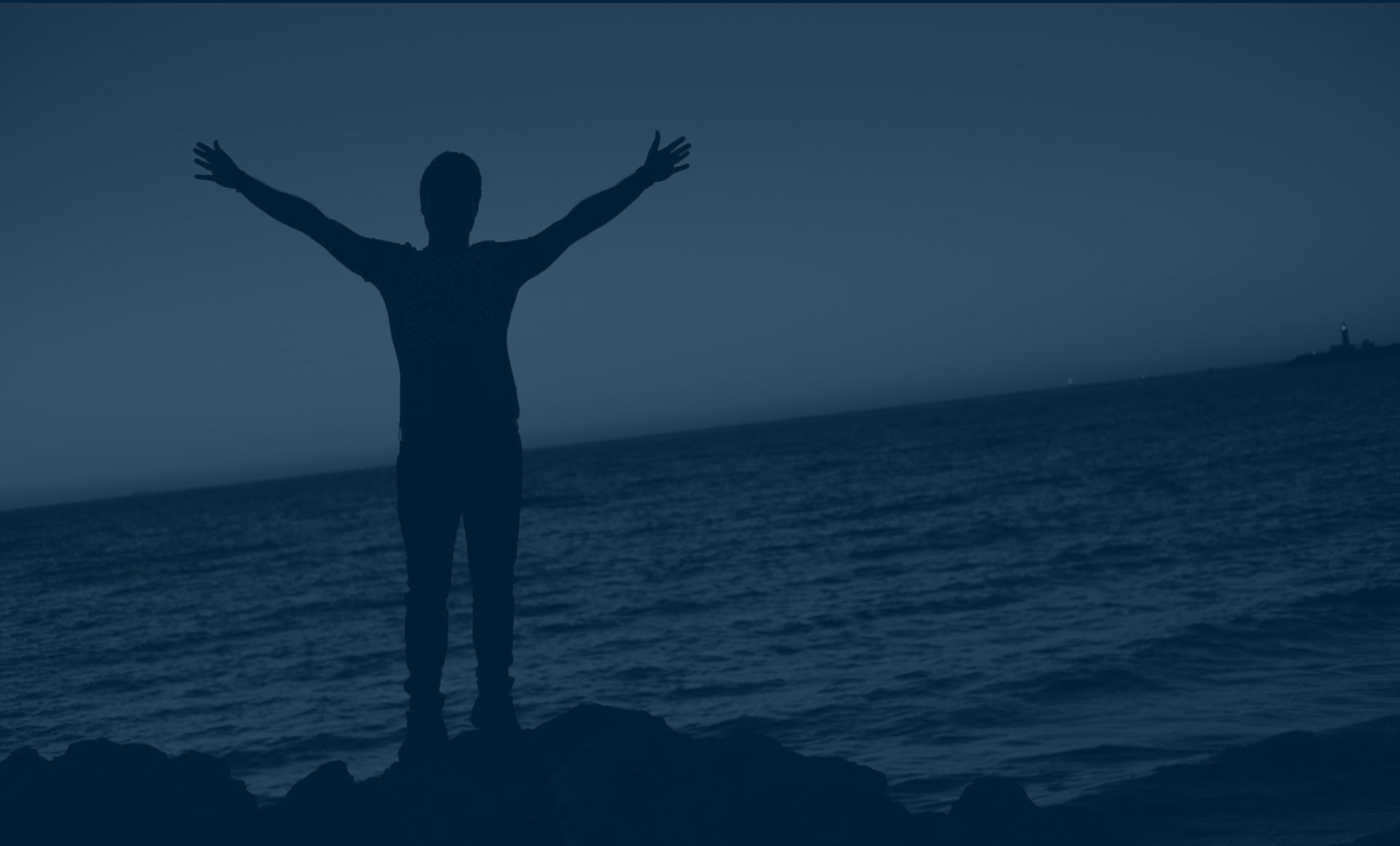 Silhouette of a person standing on rocks with arms outstretched, facing the ocean at dusk.