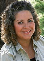 Woman with curly gray hair, smiling, wearing a tan shirt, outdoors.