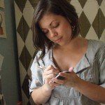 A woman with brown hair writes in a notebook. She wears a gray shirt, and a patterned wall is in the background.