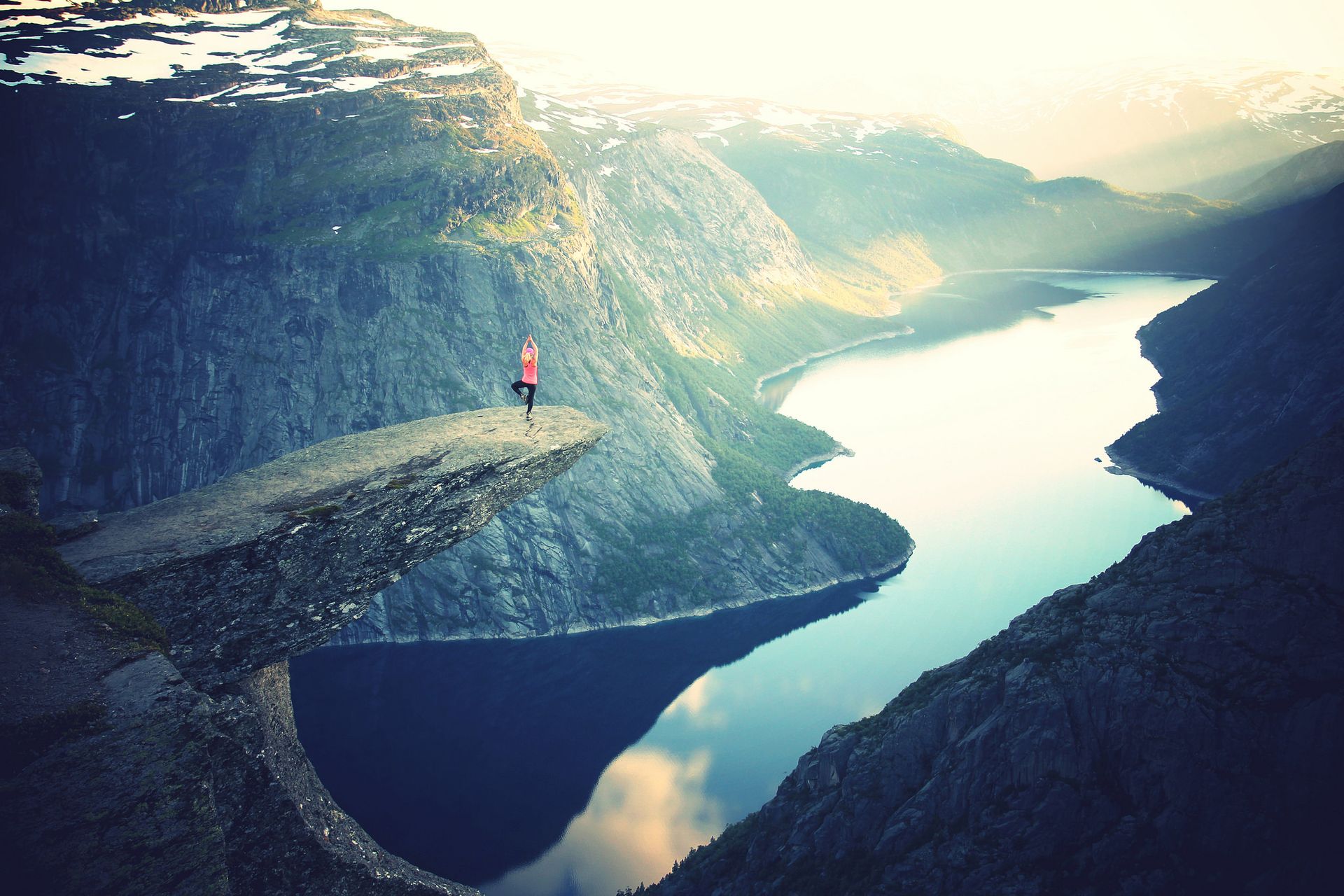 Person standing on a rock formation, overlooking a fjord. Mountains and a lake fill the background.
