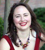 Woman with long brown hair smiles broadly, wearing a red top and beaded necklace outdoors.