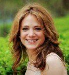 Woman with long auburn hair smiling outdoors with a blurry green background. She wears a light-colored sleeveless top.