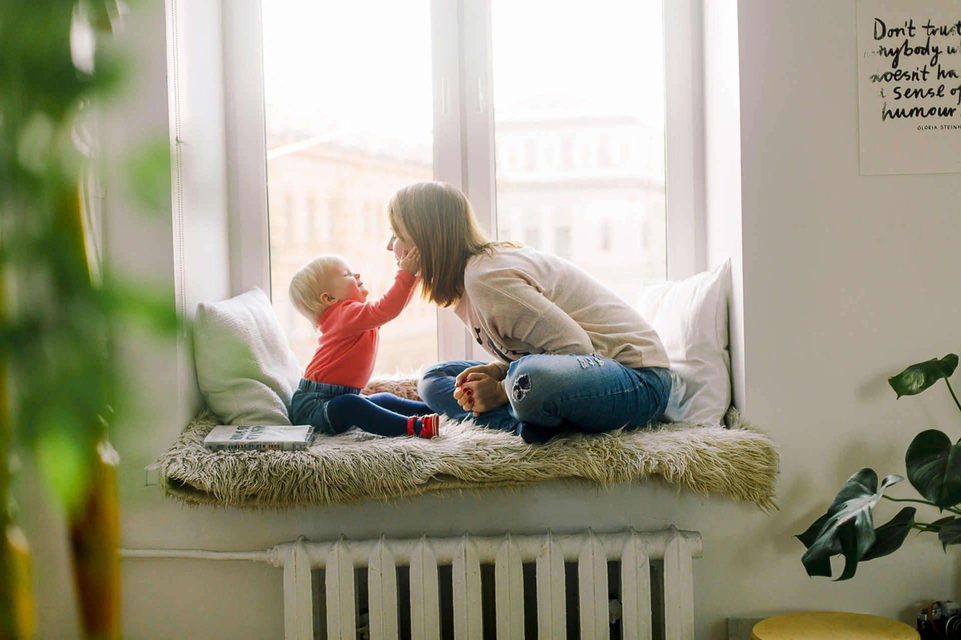 A mother and child sit on a window seat. The child touches the mother's face; both are smiling.