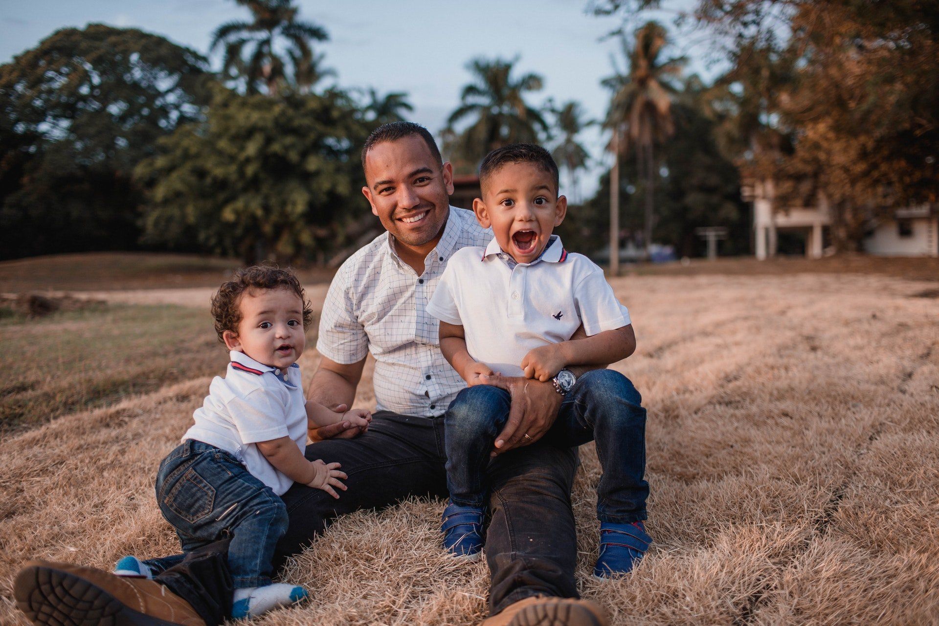 Father and two young children smiling outdoors, seated on grass. Trees and a building are in the background.