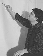A person, wearing a striped shirt, paints on a white wall with their arm outstretched, casting a shadow.