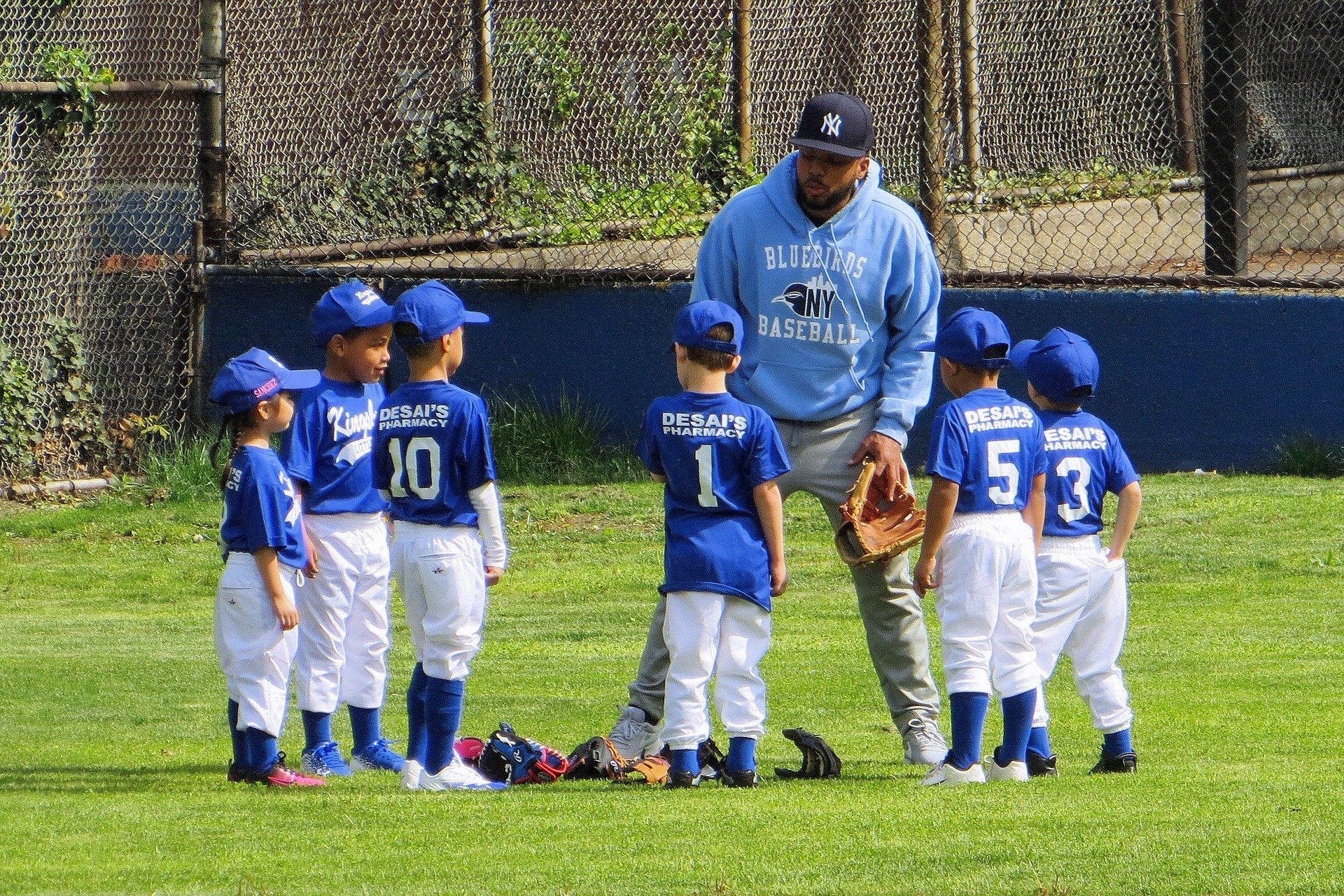 A baseball coach in a blue hoodie instructs a group of young children in baseball uniforms on a grassy field.