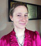 A smiling woman with short brown hair, wearing a pink top and necklace, stands indoors near a framed wall decoration.