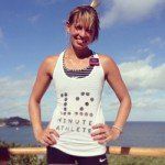 Woman in athletic attire, smiling, posing with hands on hips. She wears a white tank top with 