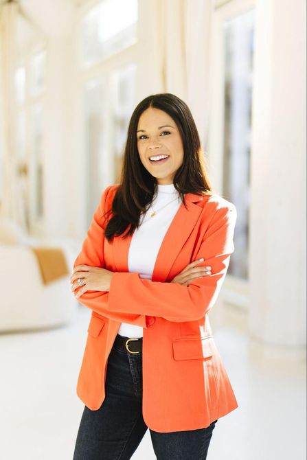 A woman in an orange jacket is standing in a hallway with her arms crossed.