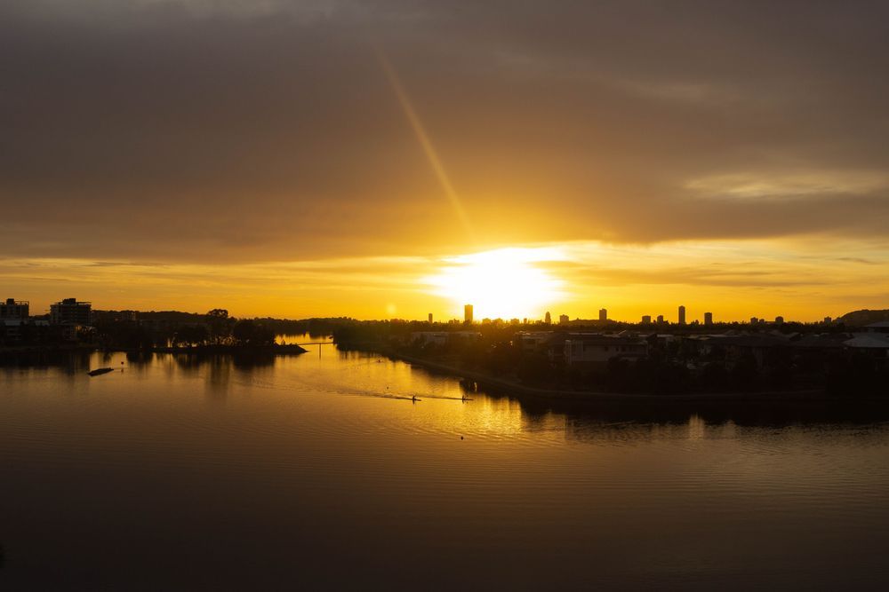 Sunset over calm water with silhouettes of buildings and trees; golden sky. — Neave Electrical in Pacific Palms, NSW